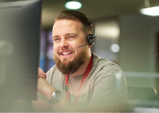 A bearded man with headphones on is looking into a computer and smiling.